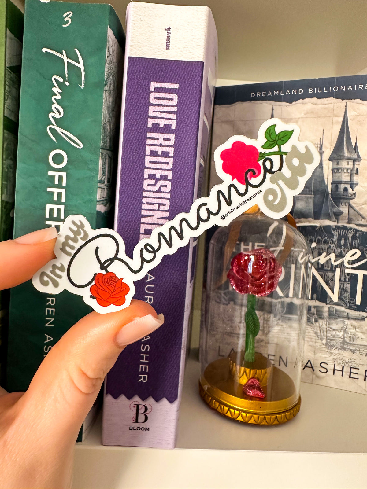 A hand holds a die-cut sticker featuring the word 'Romance' in white script over red roses, with one pink rose and green leaves on a white background. The scene is set on a bookshelf next to books including 'Fame Offer' and 'Love Redesigner'.
