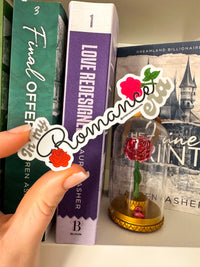 A hand holds a die-cut sticker featuring the word 'Romance' in white script over red roses, with one pink rose and green leaves on a white background. The scene is set on a bookshelf next to books including 'Fame Offer' and 'Love Redesigner'.