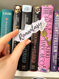 A hand holds a die-cut sticker that says 'Romantasy' in black script over a white and lavender background, positioned in front of a row of colorful fantasy book spines.