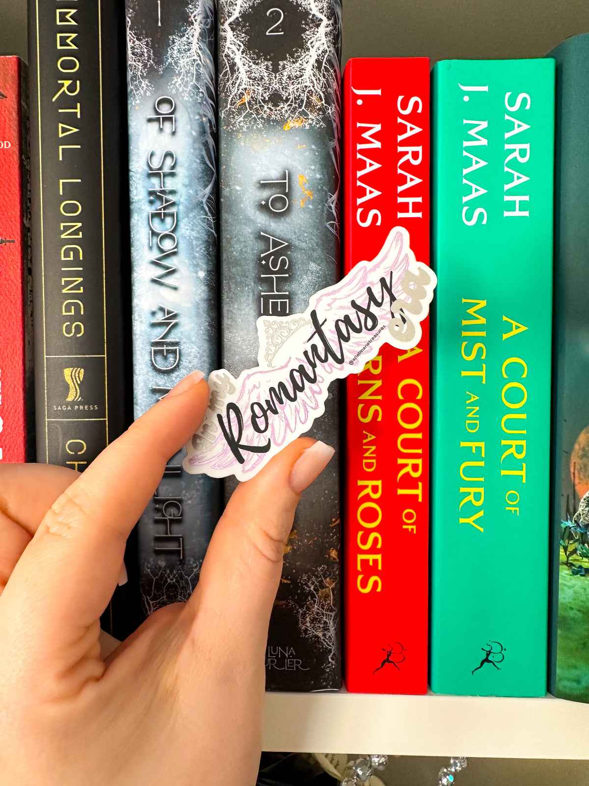 A hand holds a die-cut sticker that reads 'Romantic' in black script over a white background with a pink floral border, positioned in front of a row of colorful hardcover books on a shelf.