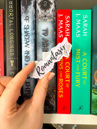 A hand holds a die-cut sticker that reads 'Romantic' in black script over a white background with a pink floral border, positioned in front of a row of colorful hardcover books on a shelf.