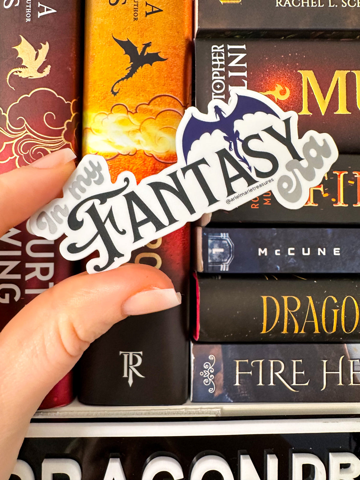 A hand holds a die-cut sticker that reads 'Fantasy' in stylized black script with a blue and white dragon illustration, positioned over a stack of fantasy book spines.