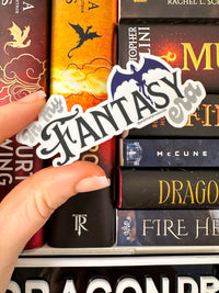 A hand holds a die-cut sticker that reads 'Fantasy' in stylized black script with a blue and white dragon illustration, positioned over a stack of fantasy book spines.