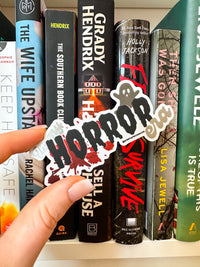 A hand holds a die-cut vinyl sticker featuring horror movie quotes like 'I HATE HORRORS' and 'I SURVIVE' in red, black, and white, with a blood splatter design. The background shows a row of horror book spines on a white shelf.