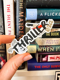 A hand holds a die-cut vinyl sticker that reads 'Thriller' in stylized black lettering with a white outline, over a stack of books.