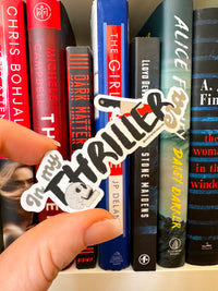 A hand holds a die-cut sticker that reads 'In My Thriller' in black script with a small white ghost illustration, positioned in front of a row of colorful book spines on a shelf.