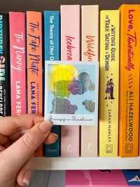 A hand holds a square glittery polaroid sticker featuring pastel clouds and the title 'Grumpy & Sunshine' in script, positioned in front of a row of colorful book spines.