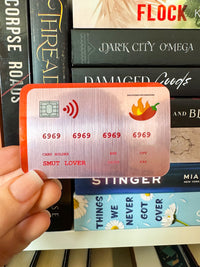 A hand holds a novelty credit card sticker with a red-orange border and a brushed silver center, featuring a chili pepper logo and the text 'SMUT LOVER'. The background shows a stack of books with titles like 'Flock', 'Dark City Omega', and 'Stinger'.