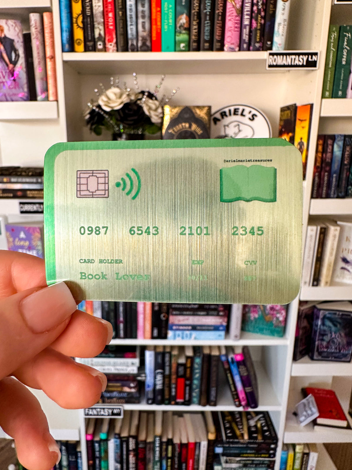 A hand holds a novelty book card sticker with a green border and gold-toned metallic background, featuring book-themed text and numbers, in front of a bookshelf filled with books.
