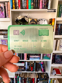 A hand holds a novelty book card sticker with a green border and gold-toned metallic background, featuring book-themed text and numbers, in front of a bookshelf filled with books.
