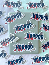 A collection of die-cut stickers featuring the word 'HORROR' in a stylized blue font with a small gray cartoon ghost and red blood splatter details, shown on a light mint green rectangular card over a white fabric background.
