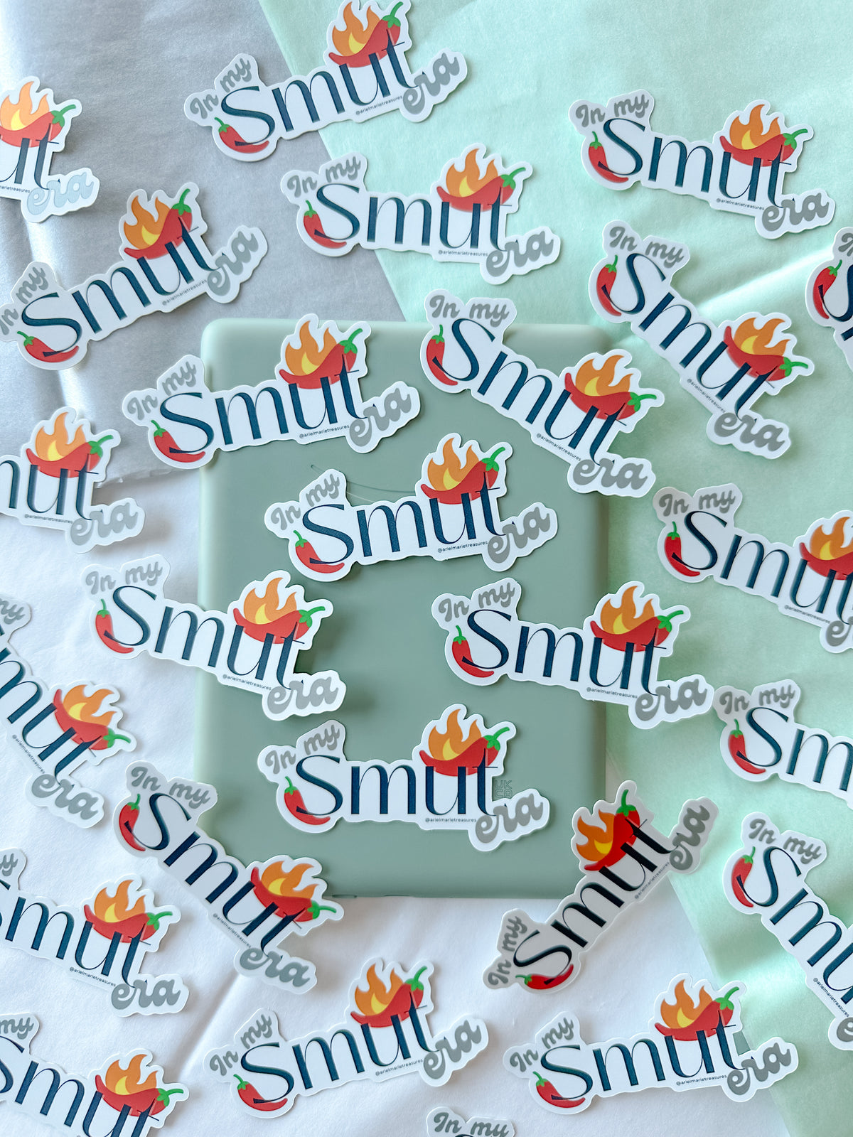 Multiple die-cut stickers with the phrase "in my Smut era" in white script and a red-orange flame chili pepper graphic, arranged on a mint green box over a white and pale green fabric background.