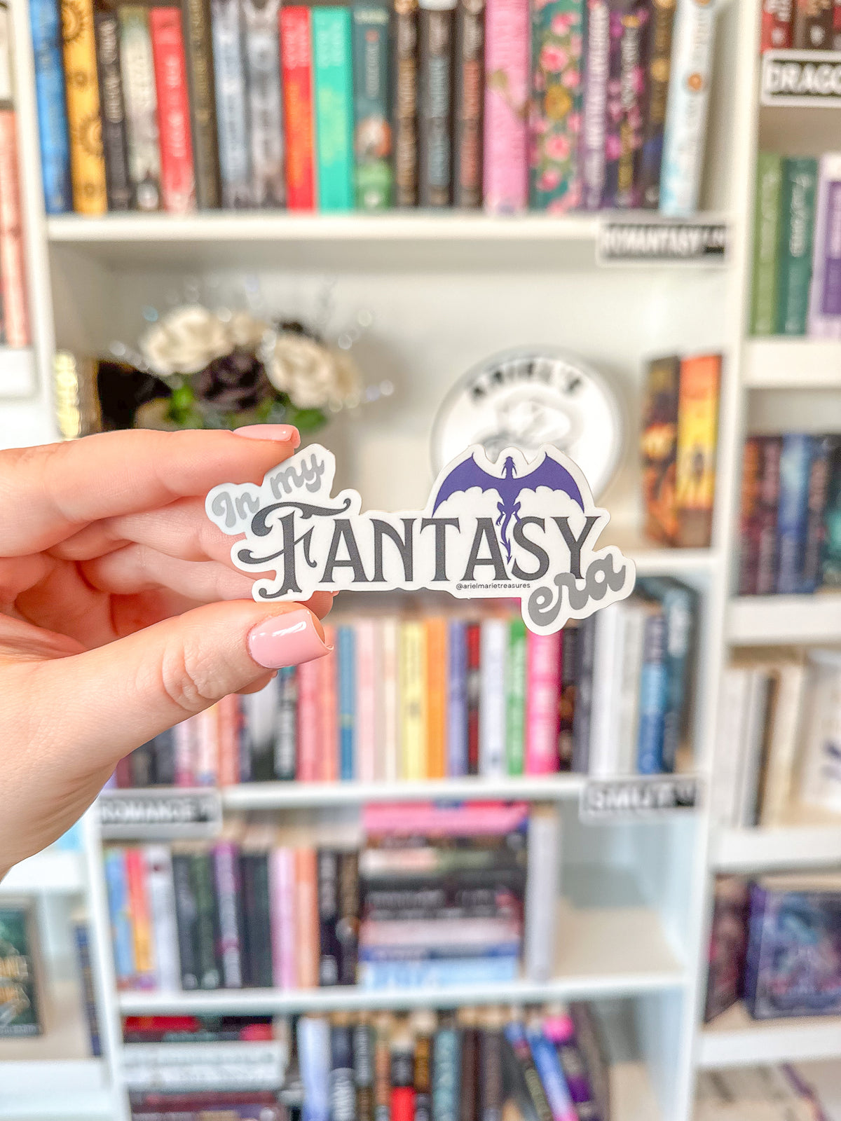A hand with pink manicured nails holds a die-cut sticker that reads 'In My Fantasy Era' with a purple dragon illustration, set against a blurred bookshelf background.