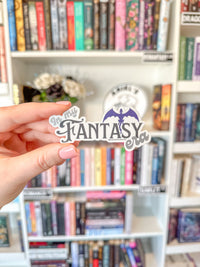 A hand with pink manicured nails holds a die-cut sticker that reads 'In My Fantasy Era' with a purple dragon illustration, set against a blurred bookshelf background.