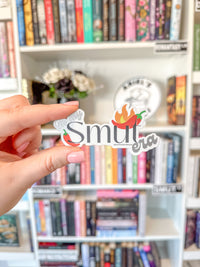 A hand holds a die-cut sticker that reads 'Smut era' with small chili pepper illustrations, in front of a blurred bookshelf background.