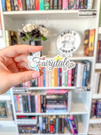 A hand holds a die-cut sticker that reads 'Fairytale' in cursive with a small winged character and crown, in front of a blurred bookshelf.