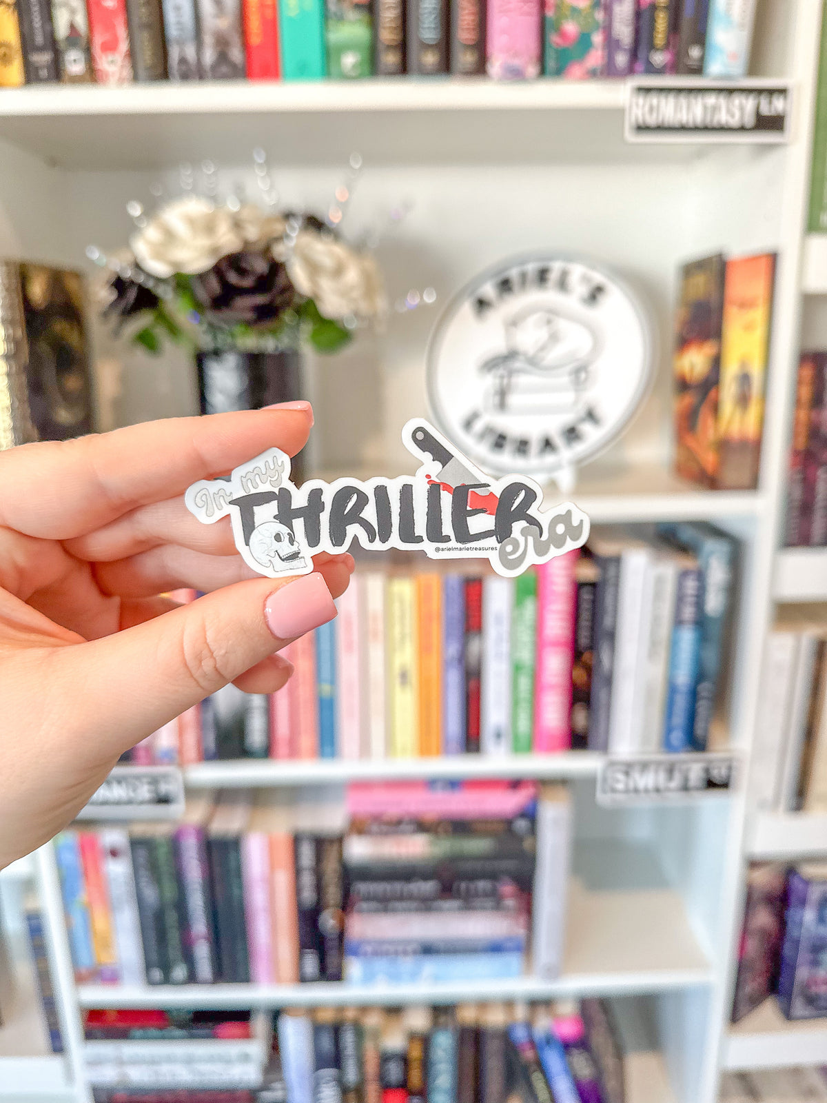 A hand holds a die-cut sticker that reads 'Thriller' with a knife graphic, in front of a blurred bookshelf with a round 'Angel's Library' sign in the background.