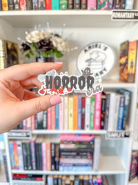 A hand holds a die-cut sticker that reads 'HORROR era' with a ghost and red splatter details, set against a blurred background of a bookshelf filled with romance novels.