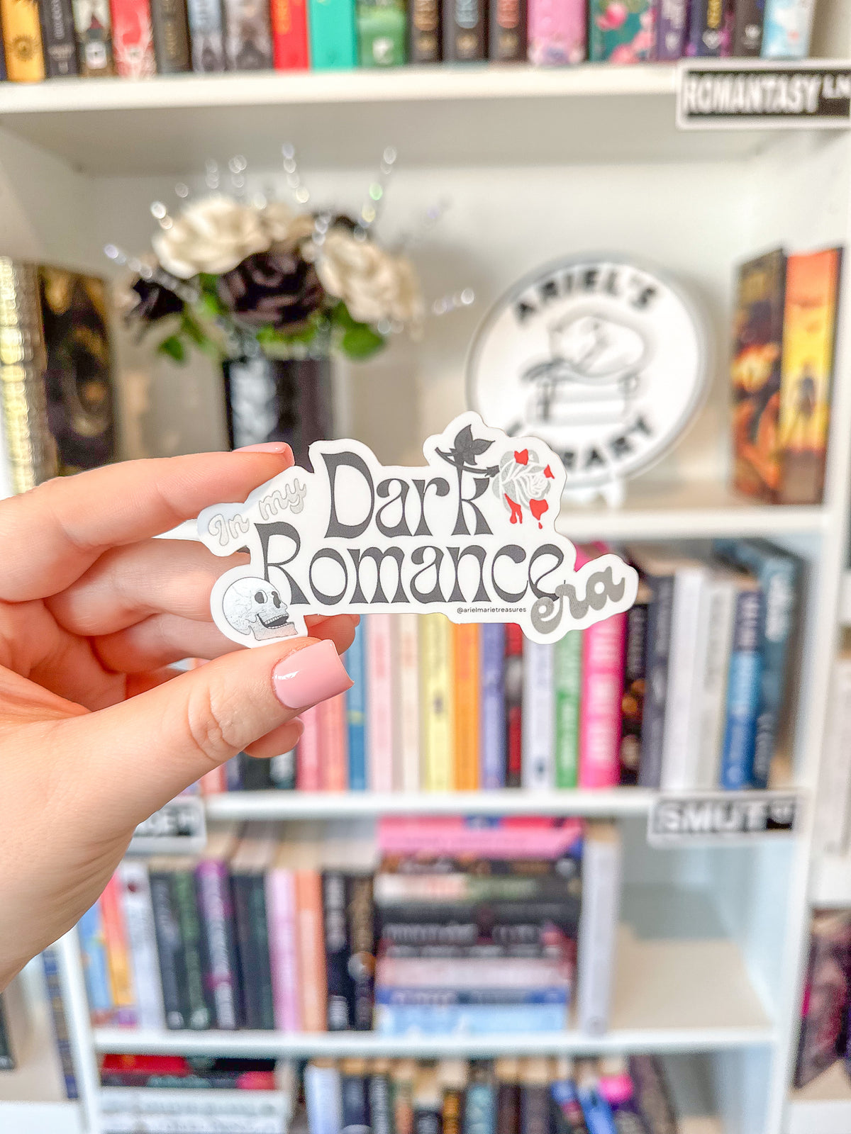 A hand with pink nail polish holds a die-cut sticker that reads 'Dark Romance' with a vampire and roses, set against a blurred background of a bookshelf.