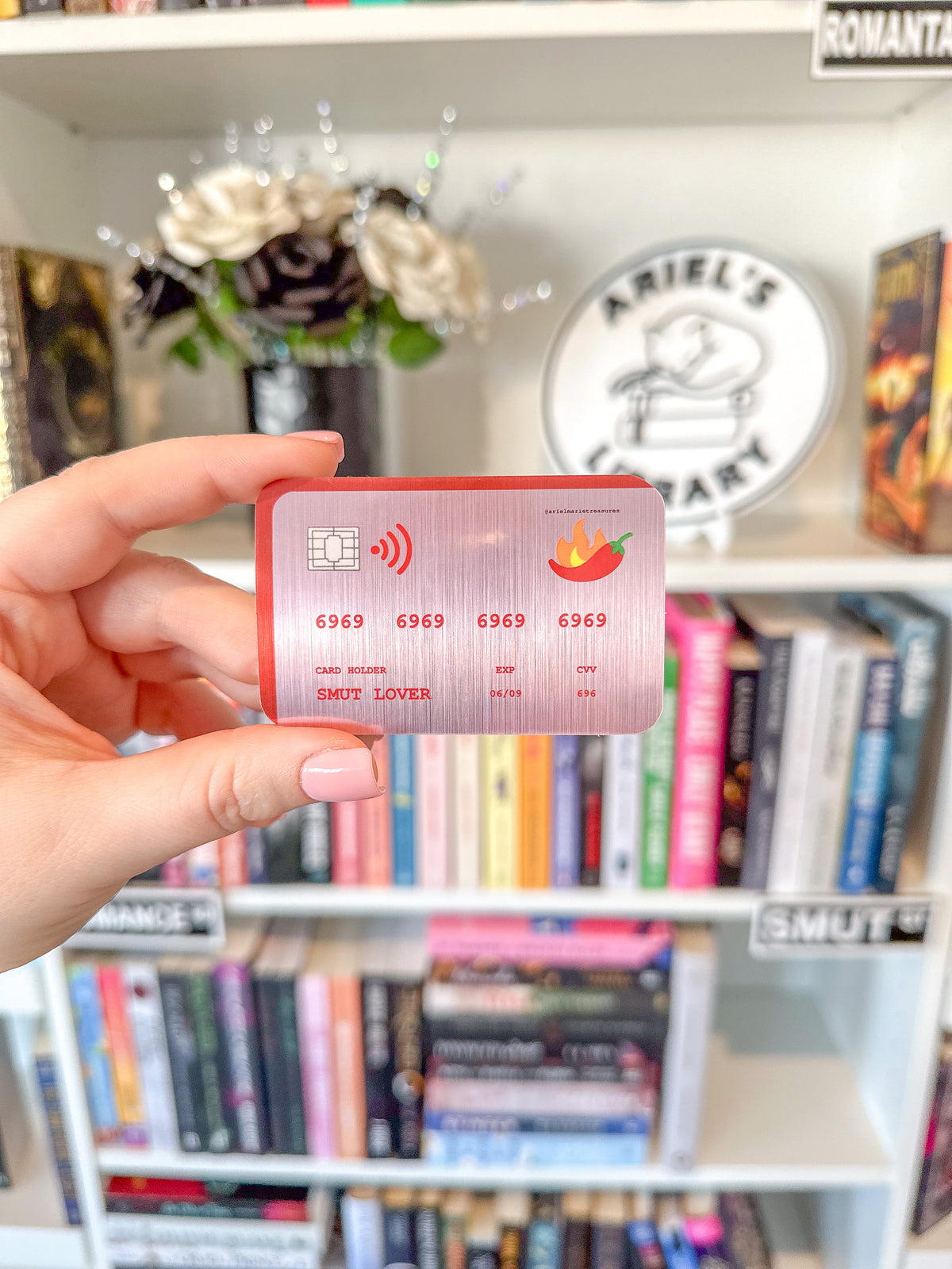 A hand holding a novelty credit card sticker with a red border and a pink metallic center, featuring placeholder card details and a chili pepper logo, set against a blurred bookshelf background.