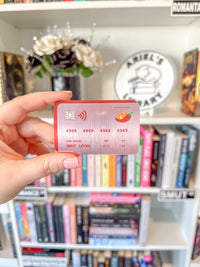 A hand holding a novelty credit card sticker with a red border and a pink metallic center, featuring placeholder card details and a chili pepper logo, set against a blurred bookshelf background.