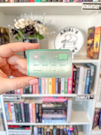 A hand holding a metallic green card sticker with a brushed silver texture, featuring placeholder credit card details and a small QR code, set against a blurred background of bookshelves.