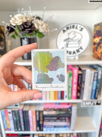 A hand holds a small square Polaroid sticker featuring glittery cloud illustrations in yellow, green, and purple tones, with the text 'Grumpy Sunshine' at the bottom. The background shows a blurred bookshelf with books, a decorative sign reading 'Ariel's Library,' and flowers.