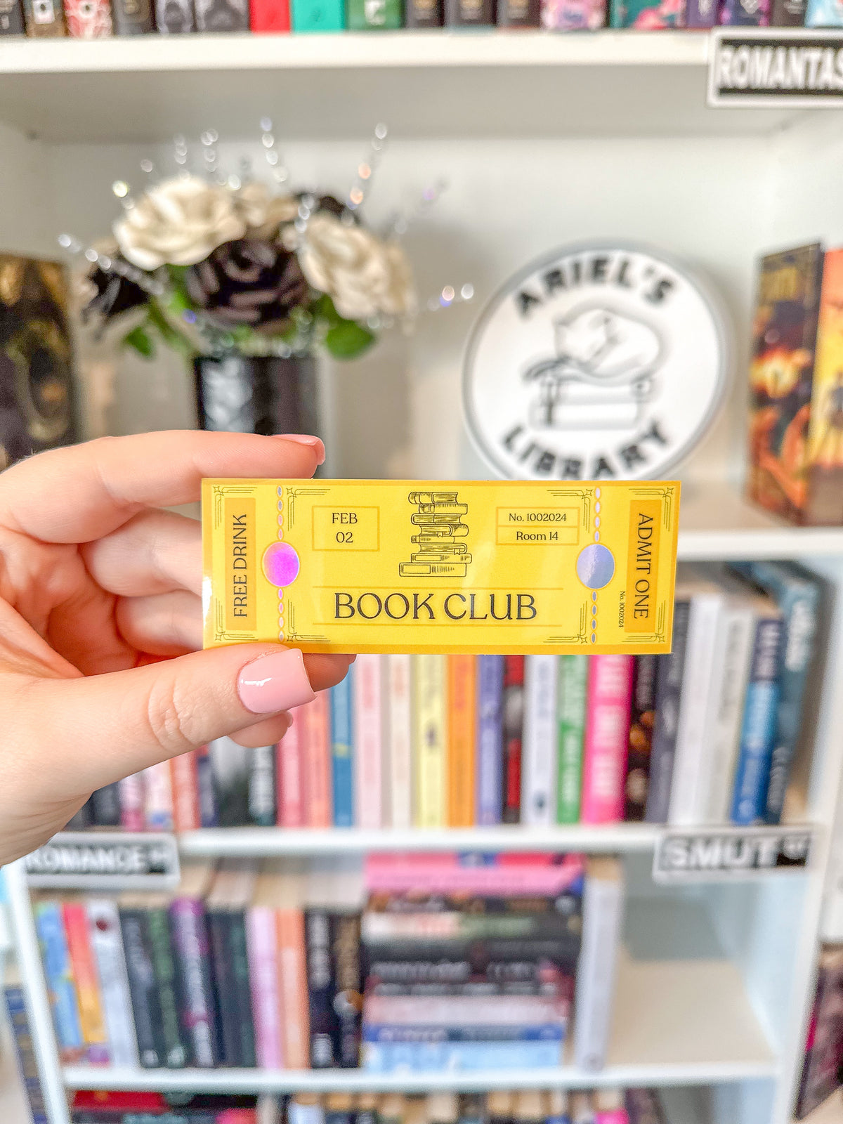 A hand with pink manicured nails holds a yellow holographic book club ticket sticker in front of a blurred bookshelf background with a round 'Ariel's Library' sign.