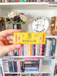 A hand with pink manicured nails holds a yellow holographic book club ticket sticker in front of a blurred bookshelf background with a round 'Ariel's Library' sign.