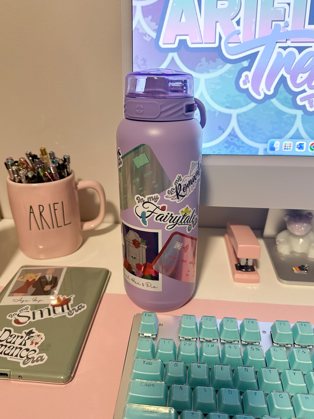 A lavender purple water bottle with a purple lid, decorated with 'The Fairytale' graphic, displayed on a desk next to a pink mug, keyboard, and other desk accessories.
