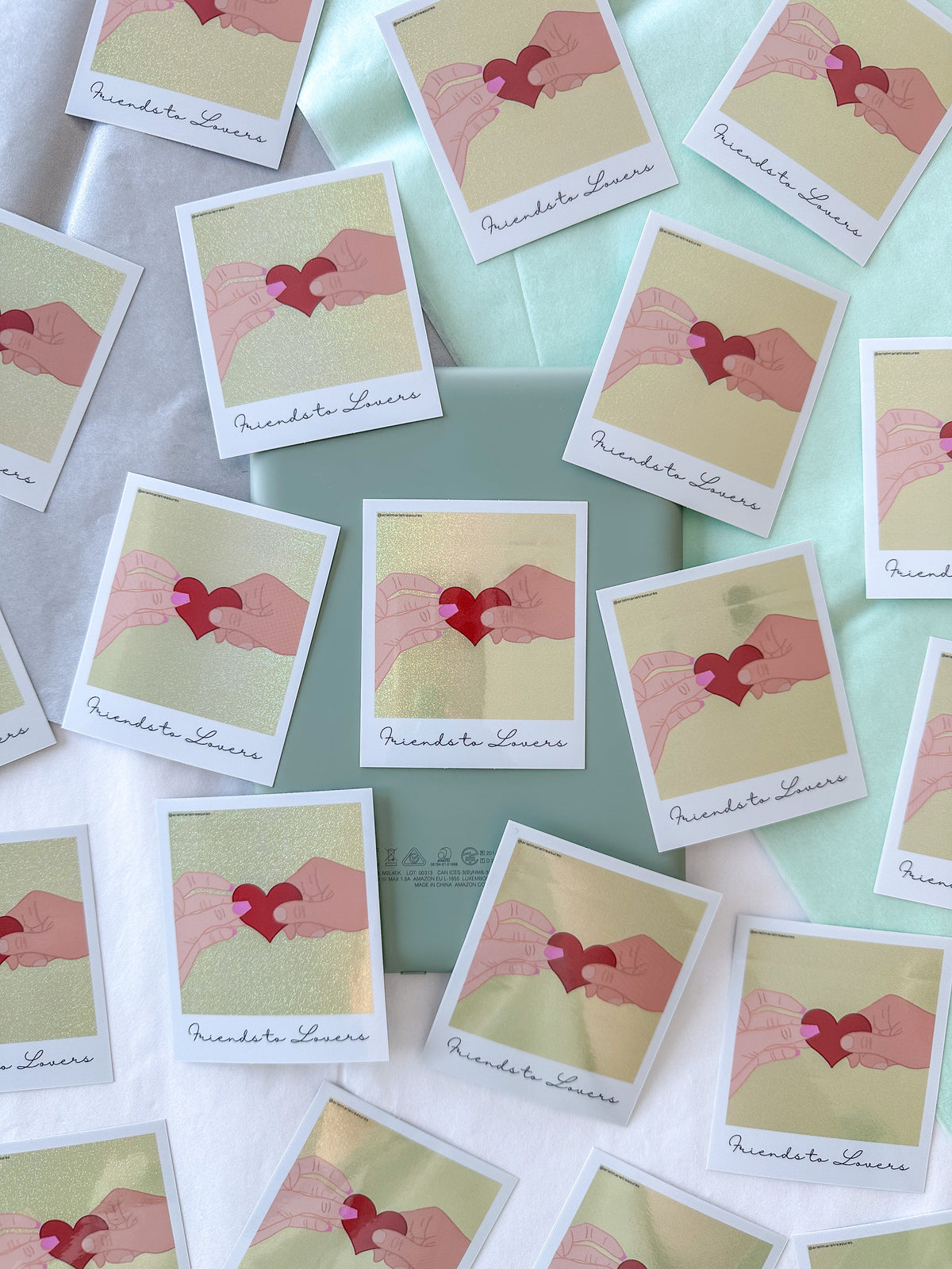 A styled flat-lay of multiple square stickers featuring an illustration of two hands holding a red heart, with the name 'Friends to Lovers' printed at the bottom. The cards are arranged on a kindle and white and mint green fabric background.