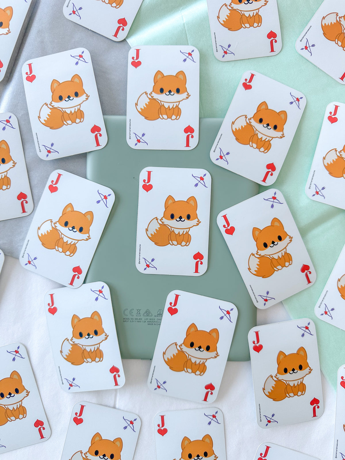 A scattered arrangement of playing cards stickers featuring a cute orange fox illustration on a white card face, photographed on a pale mint-green and white surface.