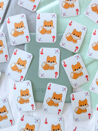 A scattered arrangement of playing cards stickers featuring a cute orange fox illustration on a white card face, photographed on a pale mint-green and white surface.