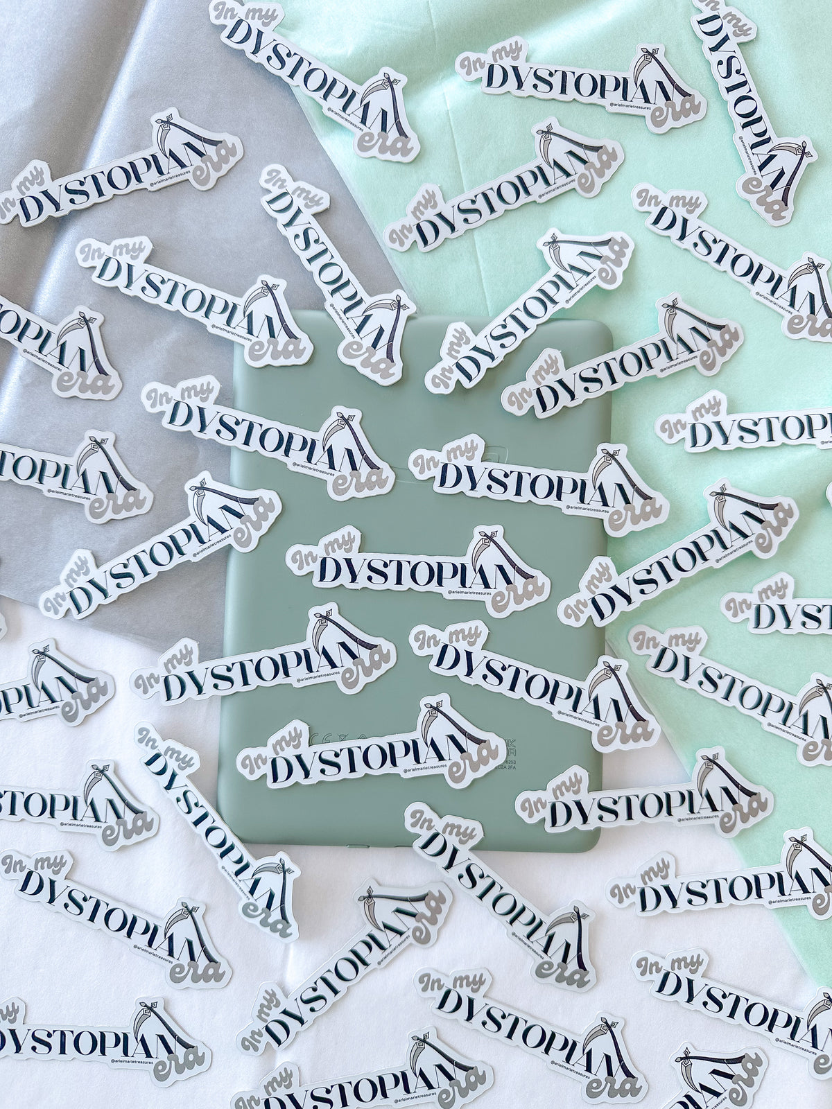 Multiple identical die-cut stickers featuring the word 'DYSTOPIA' in black script with a small black-and-white illustration, scattered across white and mint green fabric.