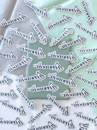Multiple identical die-cut stickers featuring the word 'DYSTOPIA' in black script with a small black-and-white illustration, scattered across white and mint green fabric.