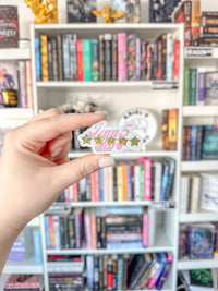 A hand holds a small die-cut sticker that reads 'I am 5 stars' in pink script with gold stars, in front of a blurred bookshelf background.