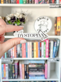 A hand with light pink manicured nails holds a die-cut vinyl sticker featuring the text 'my DYSTOPIAN life' in stylized black and white lettering. The background is a softly blurred bookshelf filled with books, creating a cozy, library-like atmosphere.