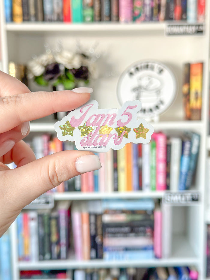 A hand with light pink manicured nails holds a die-cut sticker that reads 'I'm 5' in pink script with gold glittery stars on either side, against a blurred bookshelf background.