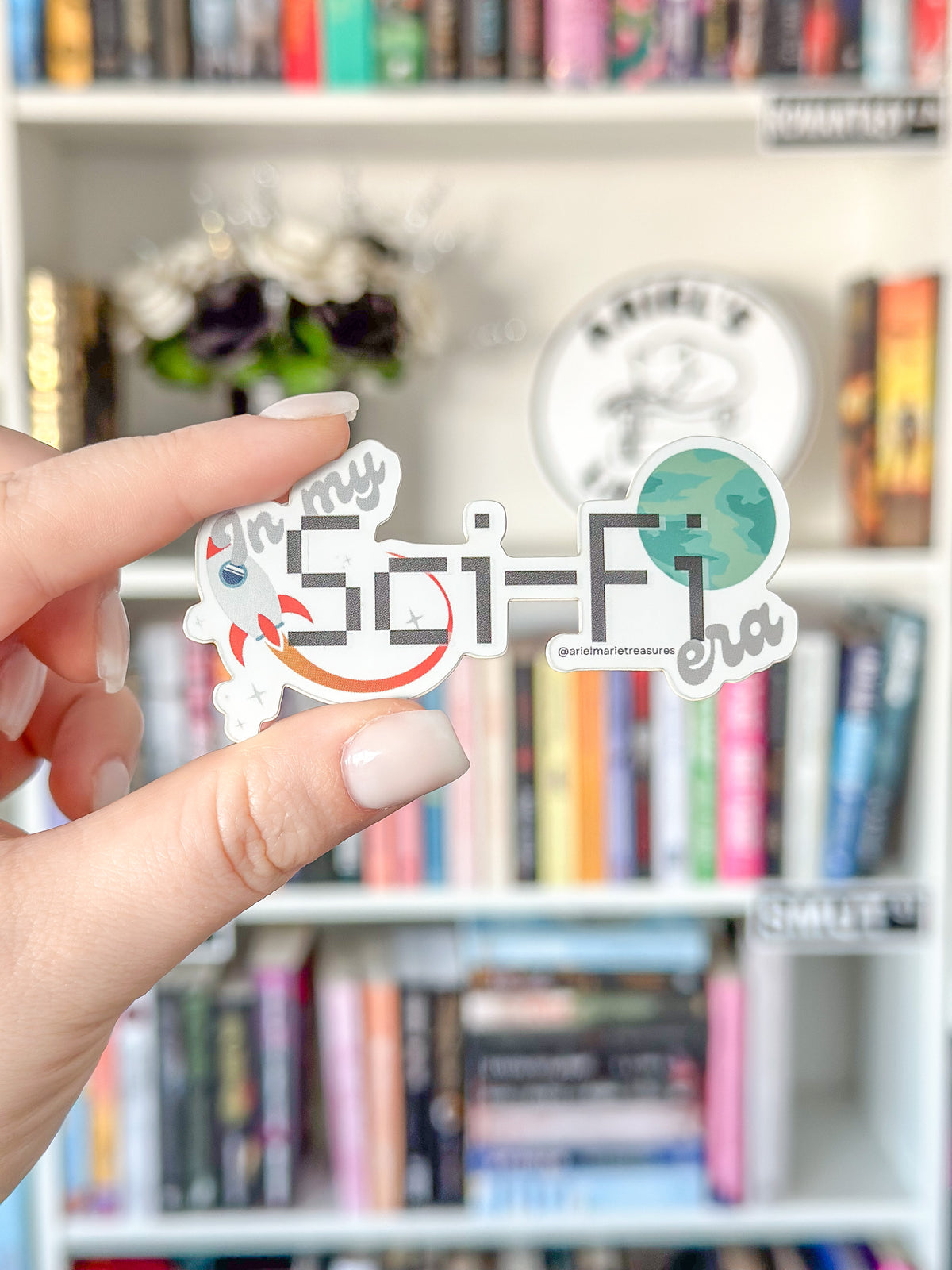 A hand with light pink manicured nails holds a die-cut sticker that reads 'in my Sci-Fi era' with a retro rocket, cat, and globe illustration. The background is a softly blurred white bookshelf filled with books.