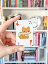 A hand holds a small white playing card featuring a cute orange cartoon fox illustration with red heart suit symbols, in front of a blurred bookshelf background.