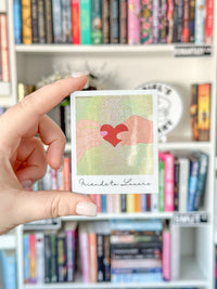 A hand holds a small square art sticker  featuring two hands forming a red heart on a glittery gold background, with the text 'Friends to Lovers' below. The background is a blurred bookshelf.