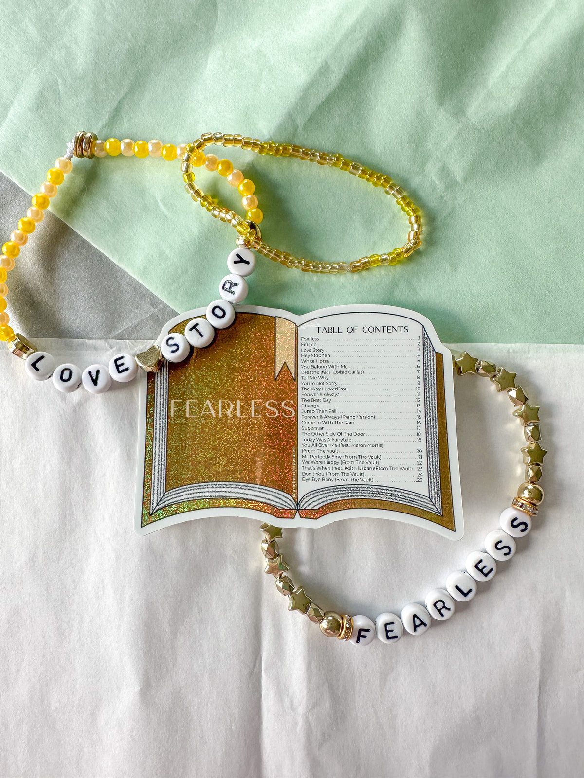 Two beaded necklaces arranged around a printed book prop on a white and mint background. One necklace is yellow beads with white letter beads spelling LOVE STORY, and the other is white beads with gold star beads and white letter beads spelling FEARLESS.