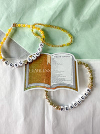 Two beaded necklaces arranged around a printed book prop on a white and mint background. One necklace is yellow beads with white letter beads spelling LOVE STORY, and the other is white beads with gold star beads and white letter beads spelling FEARLESS.