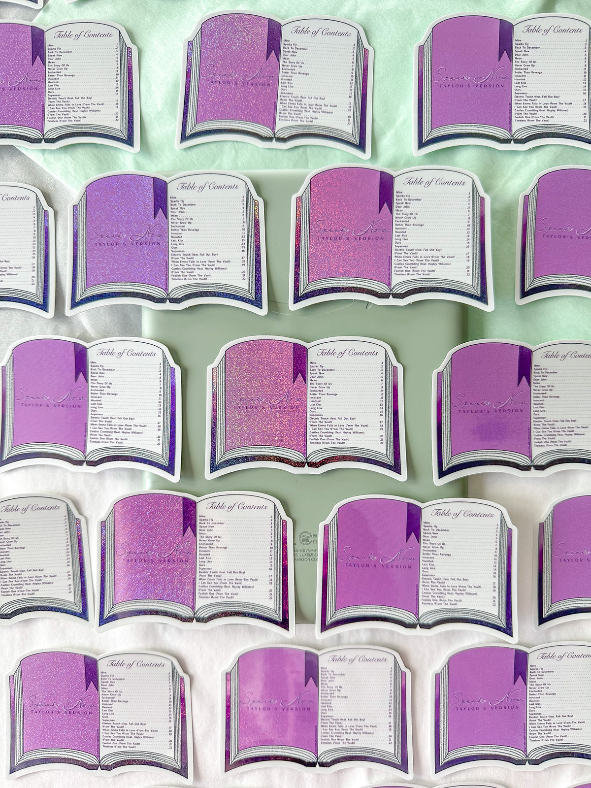 A set of book-shaped stickers arranged on a light mint green fabric background. Each book has purple glittery pages with white borders and printed text, including the heading 'Table of Contents'.