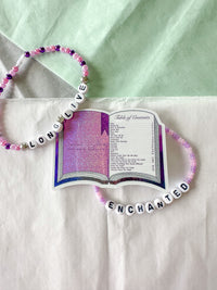 A beaded necklace with white letter beads spelling 'LONG LIVE ENCHANTED' and pink, purple, and white beads, styled on a white fabric background with a decorative open book card.
