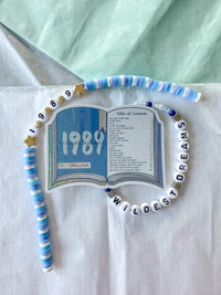 A beaded necklace and matching bracelet displayed on a white sheet of paper with a mint green fabric background. The beads are white, blue, and light blue, with some gold star accents.