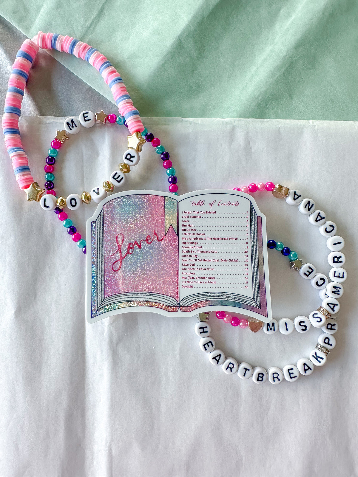 A set of beaded bracelets and a decorative book charm arranged on a white fabric surface with a pale green background. The bracelets include pink, blue, and white beads, letter beads spelling names, and one bracelet spells 'LOVE' and another reads 'ME'.