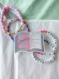 A set of beaded bracelets and a decorative book charm arranged on a white fabric surface with a pale green background. The bracelets include pink, blue, and white beads, letter beads spelling names, and one bracelet spells 'LOVE' and another reads 'ME'.