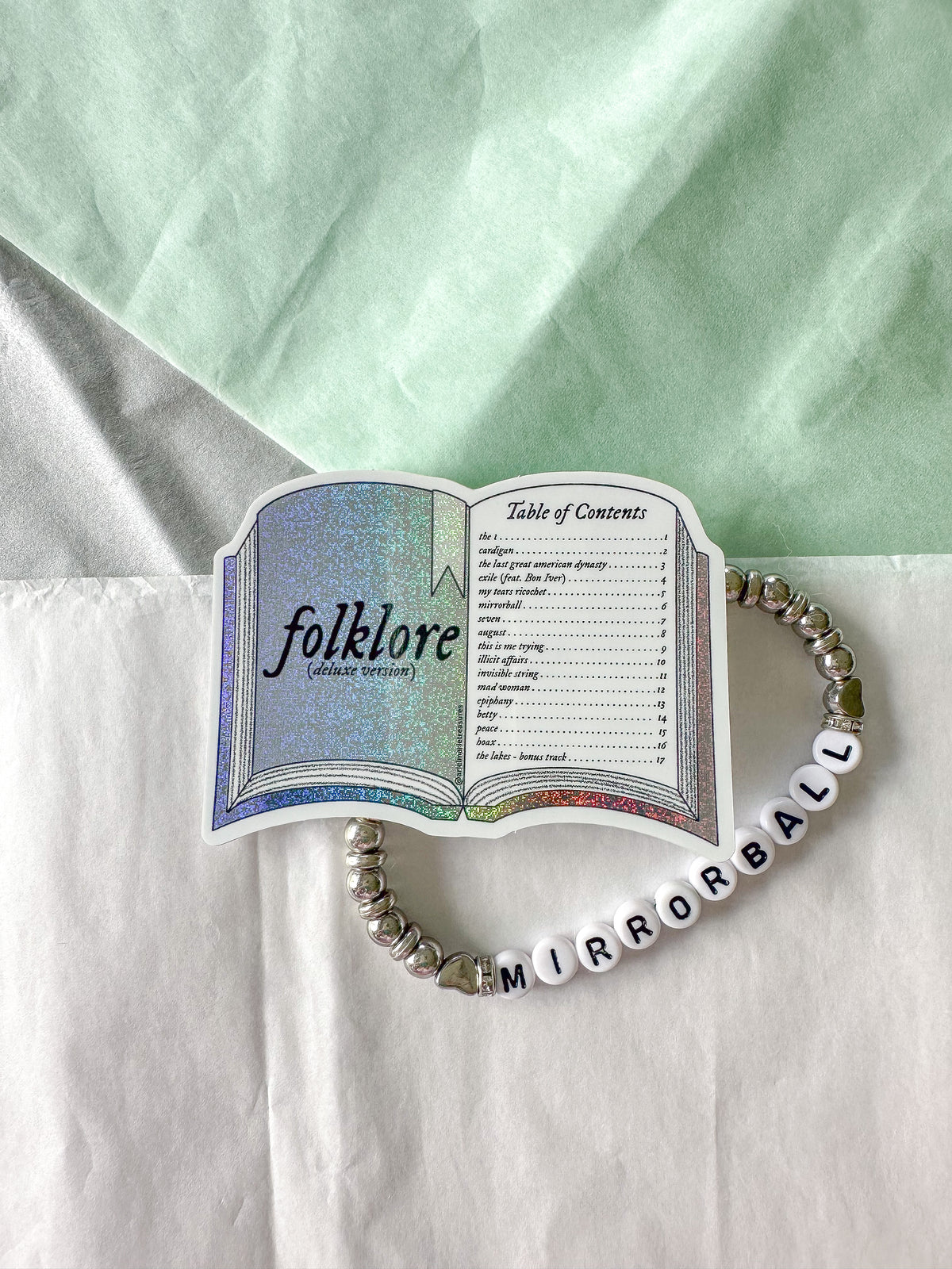 A beaded bracelet with white letter beads spelling 'MIRRORBALL' and silver spacer beads, styled on a white surface with a mint green fabric background. The bracelet is shaped like an open book page titled 'folklore (controversial)'.