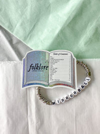A beaded bracelet with white letter beads spelling 'MIRRORBALL' and silver spacer beads, styled on a white surface with a mint green fabric background. The bracelet is shaped like an open book page titled 'folklore (controversial)'.